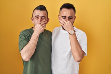 Homosexual couple standing over yellow background smelling something stinky and disgusting, intolerable smell, holding breath with fingers on nose. bad smell