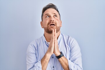 Middle age caucasian man standing over blue background begging and praying with hands together with hope expression on face very emotional and worried. begging.