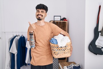 Young hispanic man with beard holding laundry basket at bedroom smiling happy and positive, thumb...