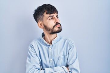 Young hispanic man with beard standing over blue background looking to the side with arms crossed convinced and confident