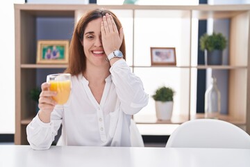 Brunette woman drinking glass of orange juice covering one eye with hand, confident smile on face and surprise emotion.