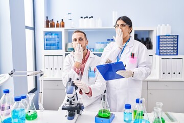 Two young people working at scientist laboratory covering mouth with hand, shocked and afraid for mistake. surprised expression