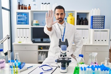 Young hispanic man with beard working at scientist laboratory doing stop sing with palm of the...