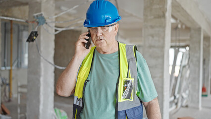 Middle age grey-haired man builder talking on smartphone with relaxed expression at construction site
