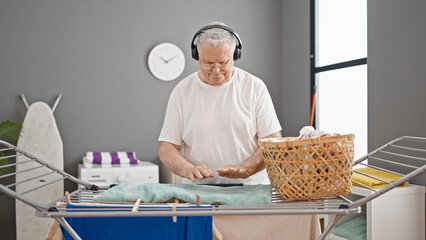 Middle age grey-haired man listening to music hanging clothes on clothesline at laundry room