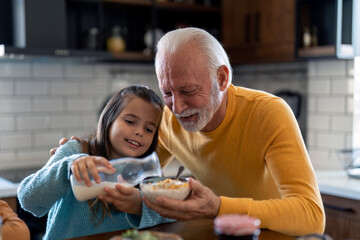 Little girl pouring milk into cornflakes cereals with grandpas help. Granddaughter and grandfather...