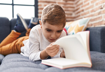 Adorable hispanic boy reading book lying on sofa at home