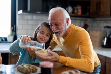 Smiling little girl with affectionate senior grandfather preparing breakfast in the kitchen at...