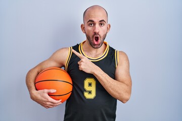 Young bald man with beard wearing basketball uniform holding ball surprised pointing with finger to the side, open mouth amazed expression.