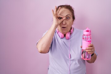 Senior woman wearing sportswear and headphones doing ok gesture shocked with surprised face, eye looking through fingers. unbelieving expression.