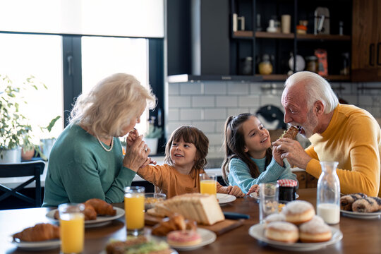 Two happy playful kids, adorable small boy and lovely little girl feeding their grandparents with homemade food, cheerful elderly couple beautiful senior woman and handsome senior man in the kitchen.