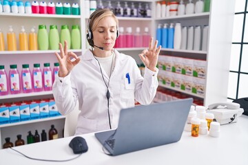 Young caucasian woman working at pharmacy drugstore using laptop relax and smiling with eyes closed doing meditation gesture with fingers. yoga concept.