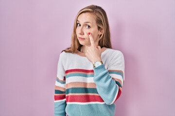Young blonde woman standing over pink background pointing to the eye watching you gesture, suspicious expression