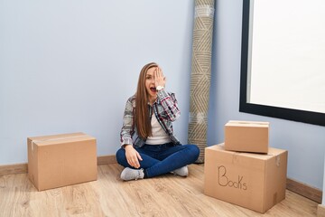 Young blonde woman sitting on the floor moving to a new home covering one eye with hand, confident smile on face and surprise emotion.