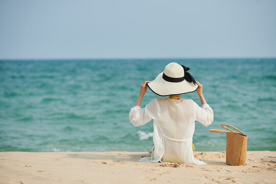 Young woman sitting on sandy beach in satin shirt and hat and looking at seascape