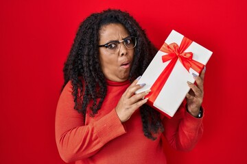 Plus size hispanic woman holding gift in shock face, looking skeptical and sarcastic, surprised with open mouth