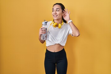 Young south asian woman wearing sportswear drinking water smiling with hand over ear listening an hearing to rumor or gossip. deafness concept.