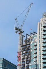 Fototapeta premium Large industrial crane machine working near tall skyscraper building, clear sky background. Modern construction industry concept