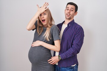 Young couple expecting a baby standing over white background surprised with hand on head for...