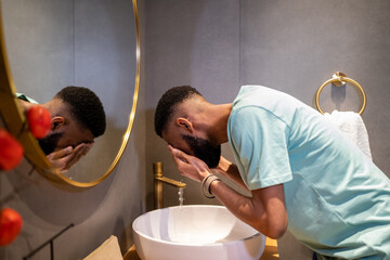 Skin care routine for men. Side view of African American millennial guy cleaning skin in morning, standing above bathroom sink and washing face with water, trying to wake up
