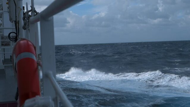 Ship in storm. High waves and splashes along side. White foam on sea. Deck railing and lifebuoy. Strong pitching. Vessel climb up wave.