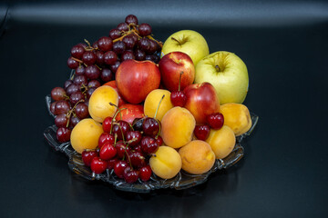A plate of fruits consisting of grapes, peaches, apricots, cherries and apples on a black background