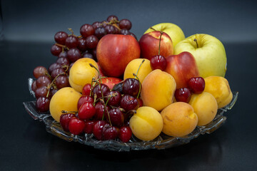 A plate of fruits consisting of grapes, peaches, apricots, cherries and apples on a black background