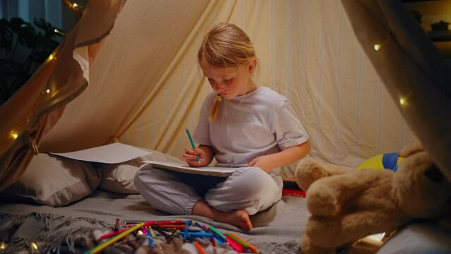 Beautiful creative little kid girl draws pictures sitting inside a play tent at home in the evening