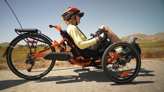 Low angle closeup of elderly senior woman riding recumbent tricycle e-bike on a path on a sunny day beside a lake.
