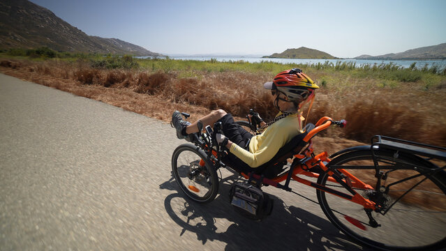 Closeup rear view of elderly senior woman riding recumbent e-bike electric tricycle bicycle on a path on a sunny day beside a lake with mountains in the distance.