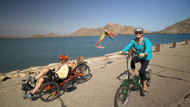 Elderly mother and mature daughter riding e-bikes beside a lake. Mother is on a recumbent electric bike.