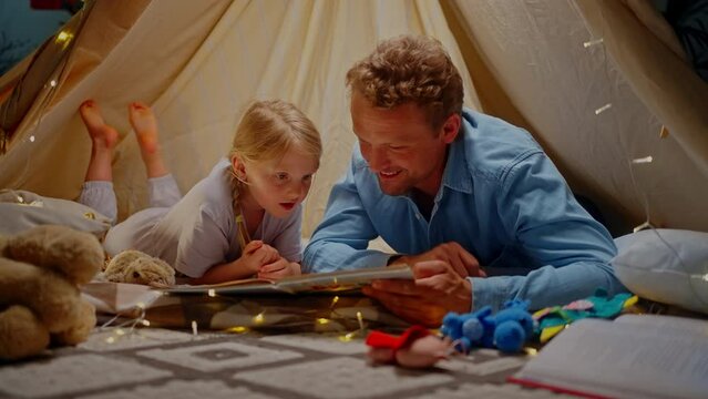 Caring loving dad and his little daughter reading fairy tales inside a play tent before going to bed
