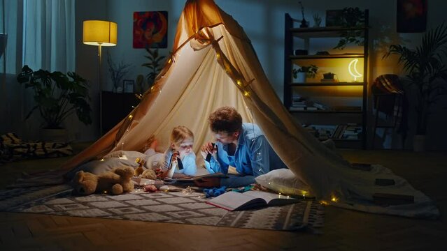 Adorable little girl and her dad read bedtime story with a flashlight, having fun inside a play tent