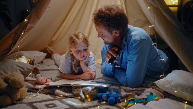 Adorable little child girl falling asleep when her father reads bedtime stories in wigwam play tent