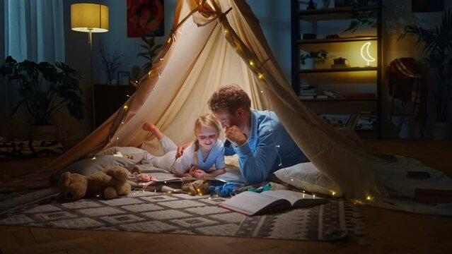 Young father and his kid daughter reading fairy tales while having fun in tent in living room