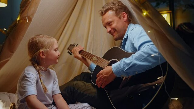 Loving young father plays guitar and sings songs with his daughter in tent illuminated with garlands