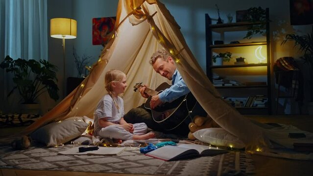 Adorable little child girl enjoys happy moments spent with her day, playing guitar in a play tent