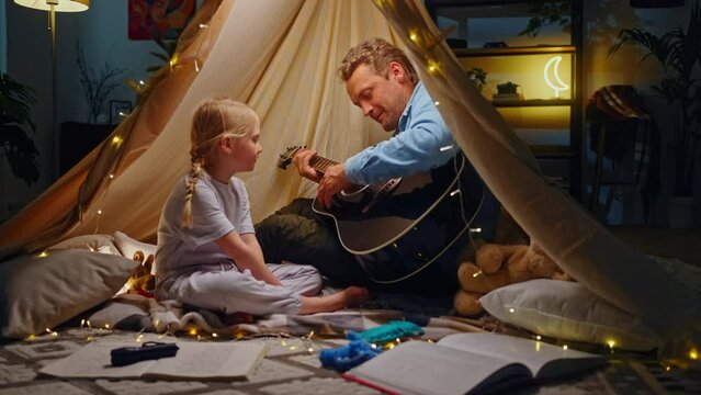 Slow motion. Happy young dad plays the guitar and sings songs with his daughter in play tent wigwam