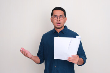 Adult Asian man showing shocked expression while holding a letter document - obrazy, fototapety, plakaty