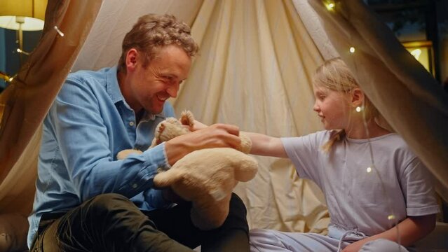 Cheerfu dad plays with his daughter in battle of plush toys, inside a tent in the light of garlands