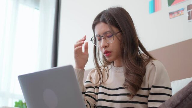 Exhausted Asian woman looks at her laptop while working overtime at home. The tired woman taking off her glasses and massaged the dry eyes. Eyestrain concept. Overwork pain. Headache from work