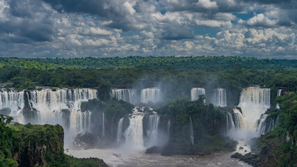 Streams of water fall in beautiful cascades.  In the bed of a stormy river, in the spray and fog, a tourist boat is visible. Lush green vegetation of the rainforest around. Iguazu Falls. Brazil.