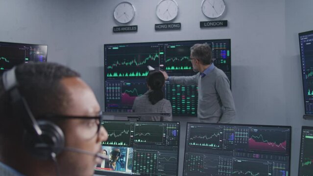 Team of financial analysts analyze exchange market charts on multiple big screens. Shot changing on African American man working at computer with displayed real-time stocks. Trading and investment.