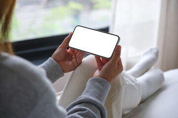 Mockup image of woman holding mobile phone with blank desktop white screen while sitting on a bed