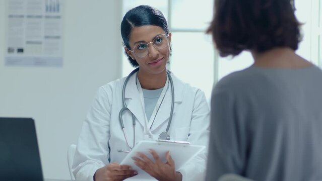 Female Doctor Taking Clipboard With Medical History, Asking Patient About Symptoms, Giving Medical Consultation In Clinic