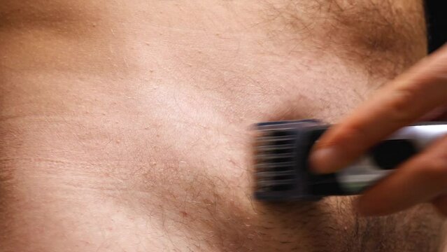 A naked man shaves his lower abdomen with an electric trimmer. Closeup. Man removes abdominal hair with an electric razor. Male beauty, personal hygiene