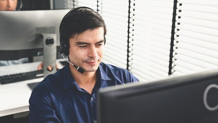 Young competent asian male call center agent working at his computer while simultaneously speaking...
