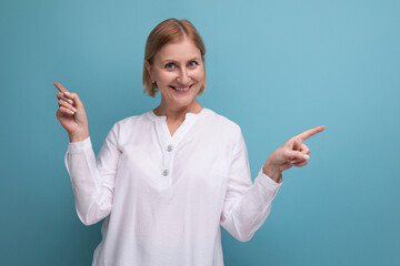 confident blond middle aged woman in white blouse pointing her finger to the side on studio background with copyspace