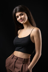 Classic studio portrait of a young brunette dressed in a black top, who is sitting on a chair against a black background.