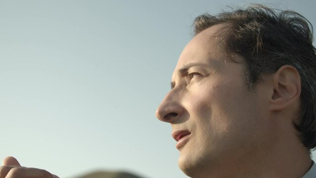 An adult man holds a dandelion seed with a parachute in his fingers and blows it off. Close up. The face is brightly lit by sunlight